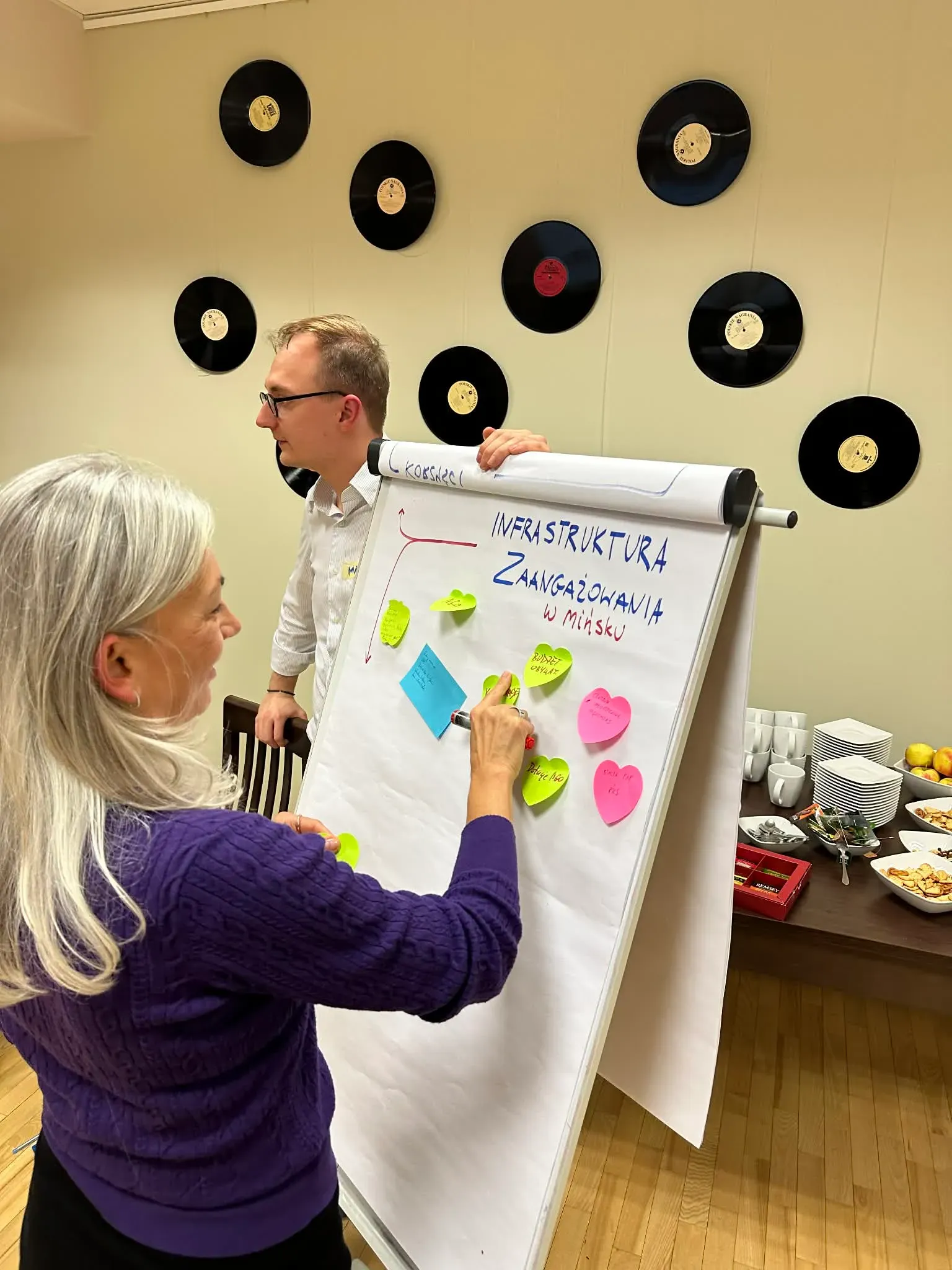 Jasne pomieszczenie. Dwie osoby przy tablicy flipchart w pokoju udekorowanym czarnymi płytami winylowymi na ścianie. Kobieta nakleja kolorowe karteczki samoprzylepne na papierze z napisem „Infrastruktura zaangażowania w Mińsku”. Na stole w tle kubki, talerze oraz przekąski.